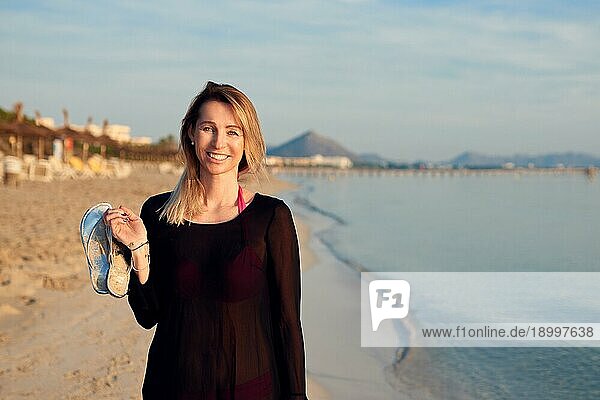 Fröhliche blonde Frau  die bei Sonnenuntergang am tropischen Strand spazieren geht