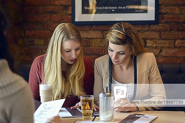 Zwei schöne blonde junge erwachsene Frauen  die an einem Stand sitzen und ihre Speisekarte in einem Restaurant mit einer Backsteinmauer hinter ihnen durchgehen