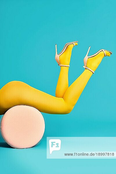 Beine der schönen jungen Frau trägt leuchtend gelbe Strumpfhosen und Schuhe mit hohen Absätzen auf Pouf auf blauem Hintergrund liegen. Entspannen  kreatives Konzept  Pop Art