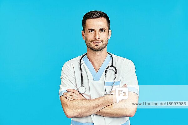 Porträt eines zuversichtlich lächelnden männlichen Arztes mit Stethoskop in medizinischer Uniform auf blauem Hintergrund mit verschränkten Armen