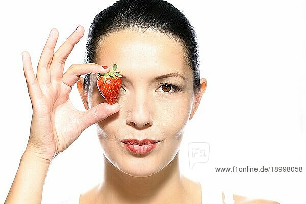 Schöne verführerische gebräunte junge Frau mit einer reifen Erdbeere  die sie an ihr Auge hält und ihre Lippen in einer sinnlichen  provokativen Weise aufreißt  während sie in die Kamera schaut  vor weißem Hintergrund