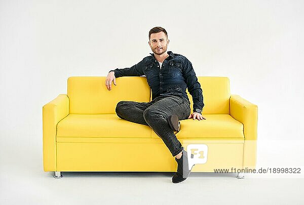 Junger trendiger Mann posiert auf gelbem Sofa sitzend und schaut in die Kamera. Entspannen Konzept