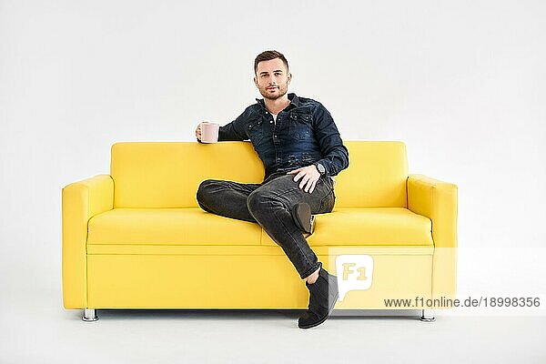 Entspannter Mann mit Kaffeetasse in den Händen sitzt auf Komfort hellen Sofa Blick zur Kamera