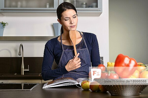 Seriöse hübsche Frau mit Schürze hält Holzkelle in der Hand  während sie am Tisch mit Obst und Gemüse in der Küche ein Rezeptbuch liest