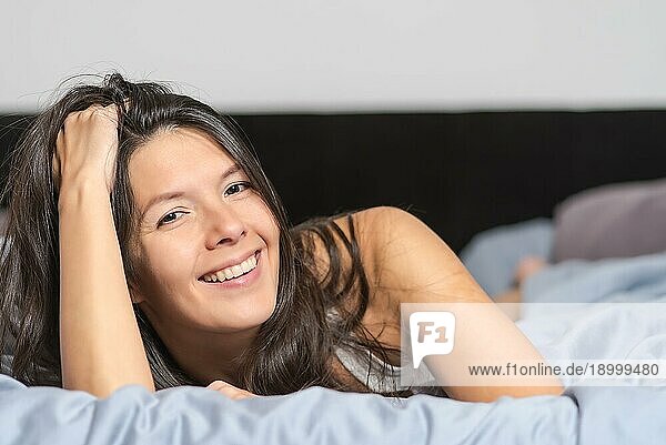 Lächelnde attraktive junge Frau  die einen faulen Tag genießt  auf dem Bauch liegend  sich in eine warme Bettdecke kuschelnd und mit einem glücklichen Lächeln in die Kamera blickend
