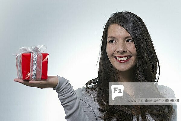 Hübsche junge Frau mit langen brünetten Haaren und einem schönen sanften Lächeln  die ein buntes rotes Geschenk für einen geliebten Menschen überreicht