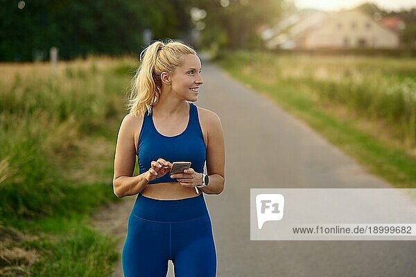 Fitte  sportliche Frau  die beim Joggen auf einem ländlichen Wanderweg ihr Handy konsultiert und dabei mit einem zufriedenen Lächeln zur Seite schaut