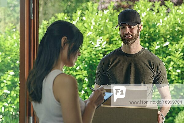Frau  die eine Lieferung von zwei Kartons mit einer Bestellung oder einem Geschenk entgegennimmt und das digitale Klemmbrett für den freundlich lächelnden Zusteller an der Türschwelle unterschreibt