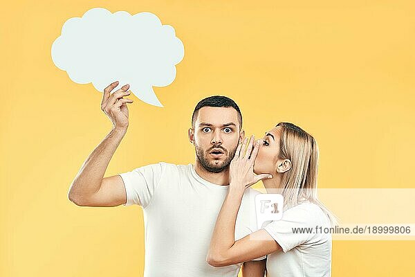 Junge Frau flüstert Geheimnisse zu Mann mit Papier Gedankenblase über gelben Hintergrund. Gossiping  Paar Beziehung Konzept