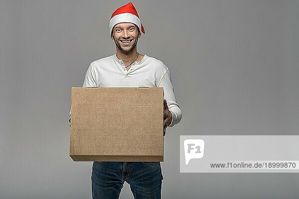 Fröhlicher attraktiver junger Mann mit Weihnachtsmannmütze  der einen großen braunen Karton für Weihnachten trägt oder ausliefert  über grauem Hintergrund mit Leerzeichen