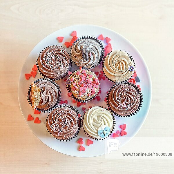 Blick von oben auf acht köstlich süße Cupcakes mit Schokoladen und Vanillecreme und verschiedenen anderen Verzierungen sowie kleinen rosa Herzchenbonbons