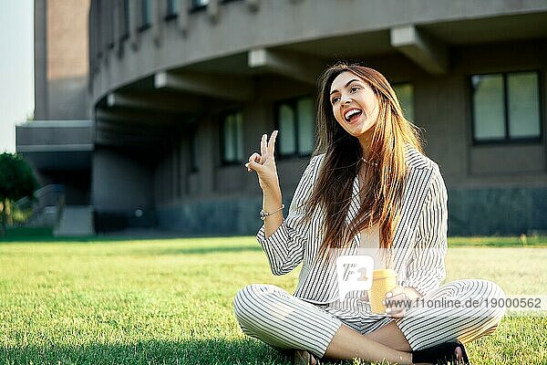Junge stilvolle Frau macht Friedenszeichen  entspannt sich mit Kaffee und sitzt auf grünem Gras. Sieg Konzept