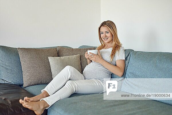 Hübsche barfuß junge schwangere Frau  die sich auf einer Couch entspannt  eine Tasse heißen Tee trinkt und glücklich in die Kamera lächelt