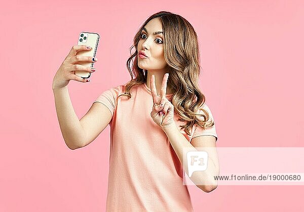 Charmante schöne Mädchen machen selfie Porträt auf Smartphone tun V Handzeichen auf rosa Hintergrund