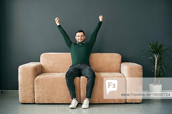 Fröhlicher junger Mann mit erhobenen Armen  der seinen Erfolg auf dem Sofa in einer modernen Wohnung feiert. Gewinnerkonzept