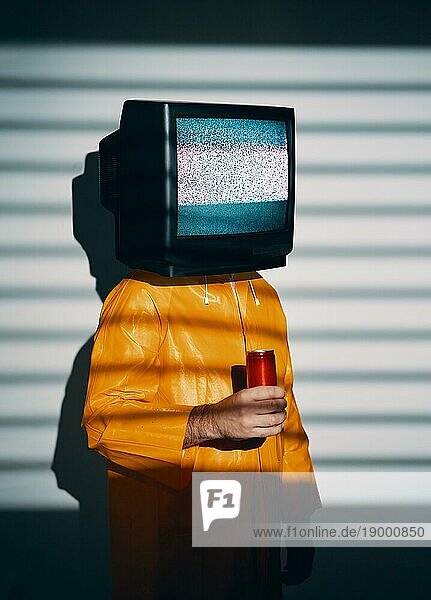 Surreale Kunst der TV süchtigen Männchen mit Fernseher statt Kopf. Medien Zombie Konzept mit männlichen in leuchtend gelben Regenmantel hält süße Soda in einer roten Dose in der Hand und posiert auf Studio in Lichter Schatten
