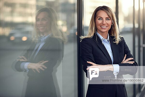 Einzelne selbstbewusste und attraktive Geschäftsfrau im blaün Anzug  die sich mit einem Grinsen an ein Fenster im Freien lehnt