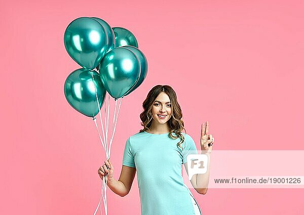 Happy hübsche Frau mit blaün Luftballons in den Händen tun Sieg Zeichen auf rosa Hintergrund