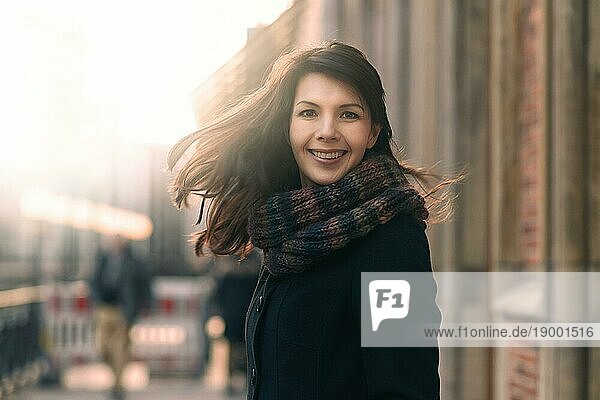 Glückliche Frau mit einem schönen Lächeln  die in ihrem Mantel und Schal auf einer städtischen Straße im Winter steht und mit einem freudigen Ausdruck in die Kamera blickt  während ihr Haar im Wind weht