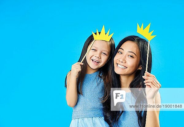Mutter und niedliche kleine Tochter mit einem Papier Krone auf Stick auf blauem Hintergrund haben Spaß zusammen. Glücklich lustige Familie Konzept