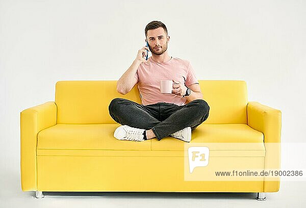 Entspannter Mann  der mit seinem Handy spricht und Kaffee trinkt  auf einem bequemen hellen Sofa sitzend