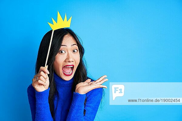 Emotionale überraschte Frau mit Papierkrone auf Stock auf blauem Hintergrund. Party  Emotionen Konzept