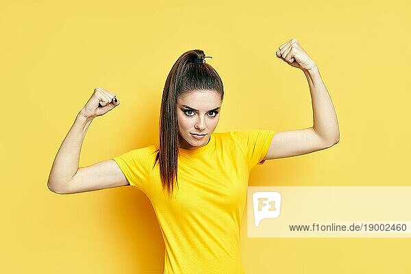 Starke  selbstbewusste junge Frau mit Armmuskeln auf gelbem Hintergrund. Frauenpower. Zuversichtlich und stolz weiblich  Erfolgspose  Fitnesskonzept