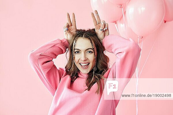 Junge hübsche Frau haben Spaß zeigen Hörner mit Fingern über den Kopf auf rosa Hintergrund. Emotionen Konzept