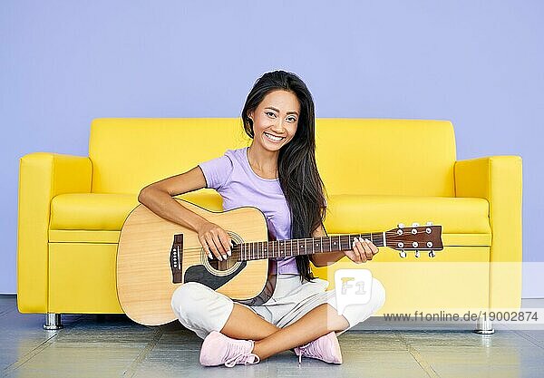 Lächelnde hübsche Frau spielt Gitarre auf dem Boden sitzend lehnt Sofa. Kreativ  Hobby  Musik Konzept