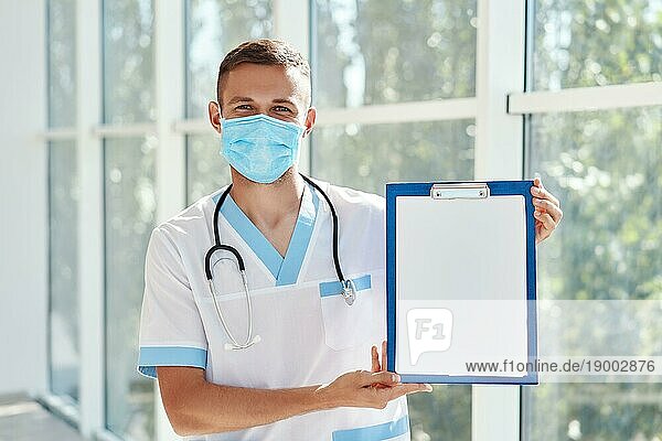 Selbstbewusster männlicher Arzt mit medizinischer Maske  der ein Klemmbrett mit einer Diagnose im Krankenhaushintergrund zeigt. Medizin  Beruf und Gesundheitswesen
