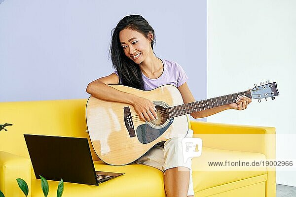 Lächelnde hübsche Frau  die lernt  Gitarre zu spielen  sieht sich eine Online Videostunde auf einem Laptop an. Onlineschulung Hobby  Musikkonzept
