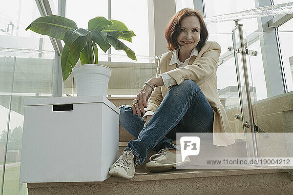 Smiling senior businesswoman sitting by box and potted plant on staircase in office