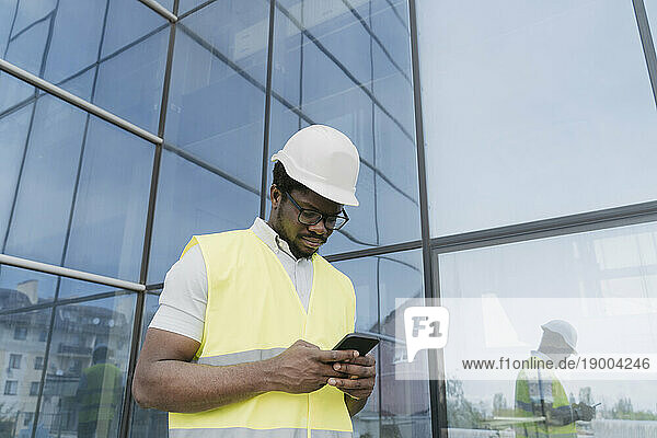 Engineer in reflective clothing text messaging on smart phone in front of glass building