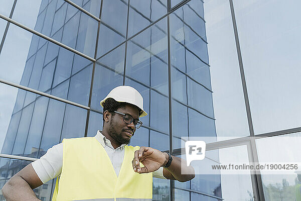 Engineer in reflective clothing checking time on wristwatch in front of glass building
