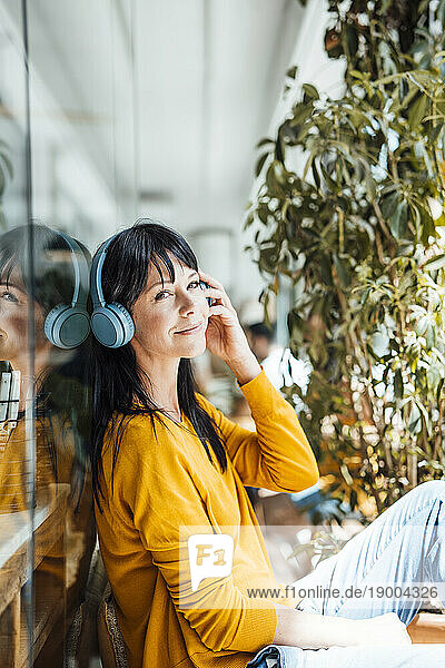 Smiling woman wearing wireless headphones listening to music by glass