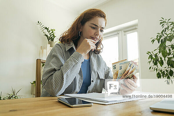 Confused young woman calculating financial bills and counting currency at home