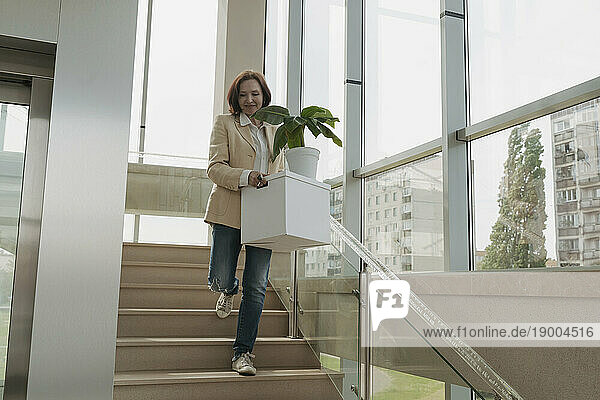 Senior businesswoman carrying box with potted plant on staircase in office