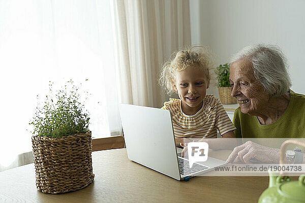 Smiling grandmother and granddaughter using laptop on table