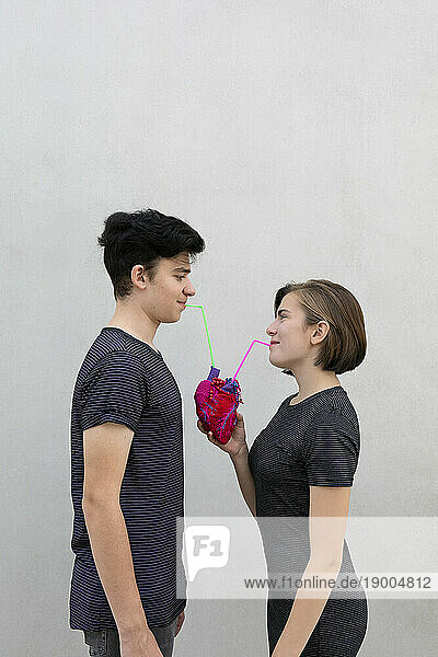 Teenage couple drinking with straw from model heart against gray background