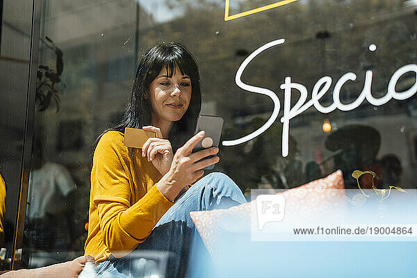 Smiling woman with credit card using mobile phone seen through glass