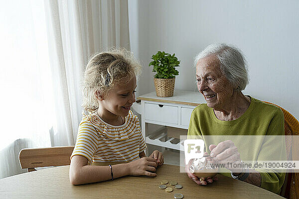 Smiling grandmother and granddaughter putting coin in piggy bank