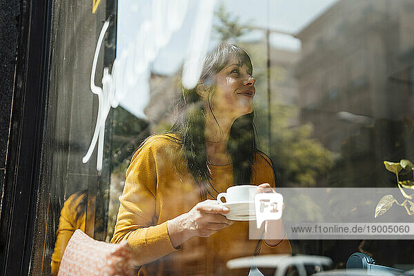Happy mature woman with coffee cup sitting in cafe seen through glass