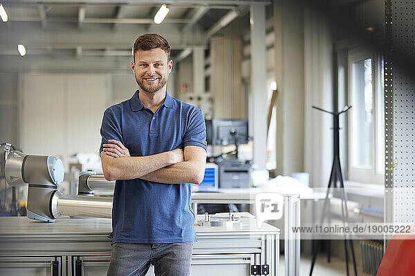 Smiling technician with arms crossed standing by table in industry