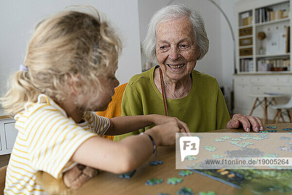 Smiling grandmother looking at granddaughter solving jigsaw puzzle