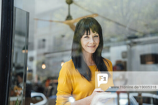 Smiling mature woman with smart phone seen through glass in cafe