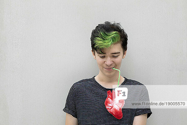 Teenage boy with dyed green hair drinking from heart against gray background