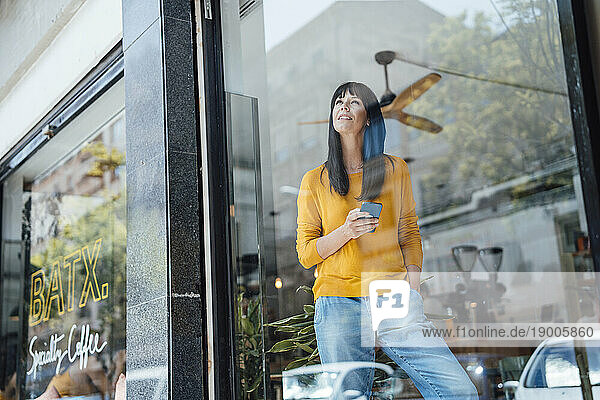 Thoughtful mature woman standing with smart phone seen through glass