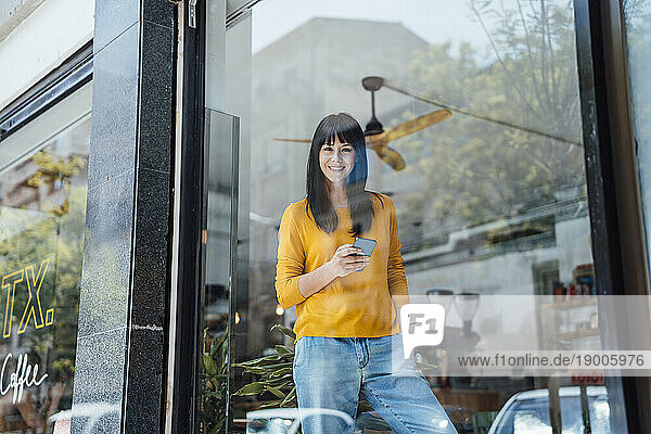 Happy woman standing with mobile phone seen through glass