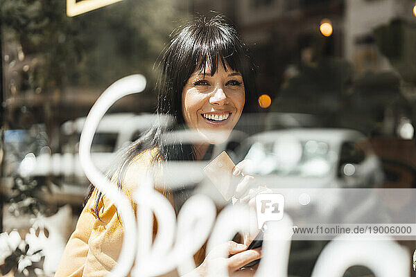 Happy mature woman with smart phone and credit card seen through glass