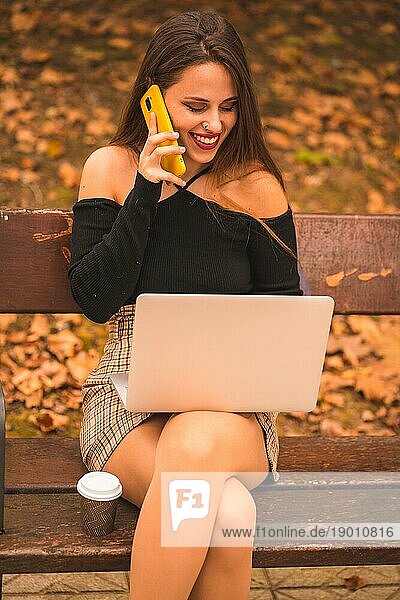 Porträt einer Frau im Herbst in einem Wald mit braunen Blättern  die am Computer sitzt und lächelnd am Telefon spricht